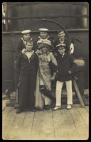 view Six sailors, one in drag, pose for a group portrait on the deck of H.M.S. St. George. Photographic postcard, 191-.