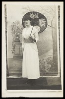 view An amateur actor in drag poses with an open parasol, in front of painted scenery. Photographic postcard, 191-.