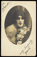 view A man in drag wears make-up and is holding flowers, posing for his portrait. Photographic postcard, ca. 1918.