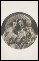 view Two young men in drag wearing Japanese costumes, pose together holding fans, underneath a parasol. Photographic postcard, 1905.