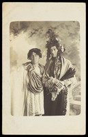 view Two men in drag, one wearing a detailed feathered head garment, pose for a portrait. Photographic postcard by F. Wood, 190-.