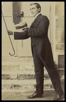 view A man wearing a morning suit holds up his cane and boater. Photographic postcard, 191-.