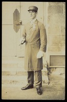 view A man dressed wearing a long coat and a hat bearing the word "Cook", stands outside a building. Photographic postcard, 191-.