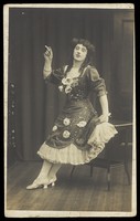 view A performer in drag, wearing a large flowery dress, poses with a cigarette on stage. Photographic postcard, ca. 1908-1910.