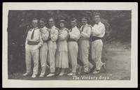 view Entertainers, one in drag, pose in a line in front of foliage. Photographic postcard, 191-.