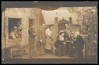view Actors, some in drag; with the attention focused on a character sitting at a table. Photographic postcard, 191-.