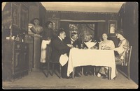 view Belgian prisoners of war, some in drag, sit at a table within a very detailed stage set at Friedberg camp, Germany. Photographic postcard, 191-.