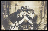 view Three actors, one in drag, pose lighting cigarettes.