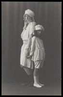 view Ralph Mellor in drag and a young actress stand back-to-back on stage. Photographic postcard by L.S. Langfier, 192-.