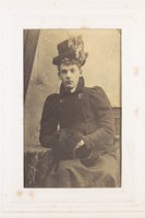 view A man in drag, seated, wearing dark clothing and a black hat. Photograph, 189-.