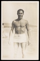 view A man stands on a beach wearing a white bathing costume. Photographic postcard, 193-.