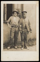 view Two soldiers stand arm-in-arm outside a building of corrugated metal (barracks?). Photographic postcard, 191-.