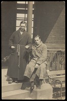 view Two men wearing dressing gowns sitting arm-in-arm on a terrace. Photographic postcard, 192-.