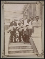 view Four men wearing aprons and uniforms standing by a trolley with half-dissected cadavers on it. Photograph.