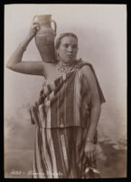 view An Algerian woman with a tattoo or henna design on her face, carrying a clay water pot against a studio backdrop.