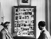 view M0005452: Two men standing in front of a display of records of cures at Lourdes